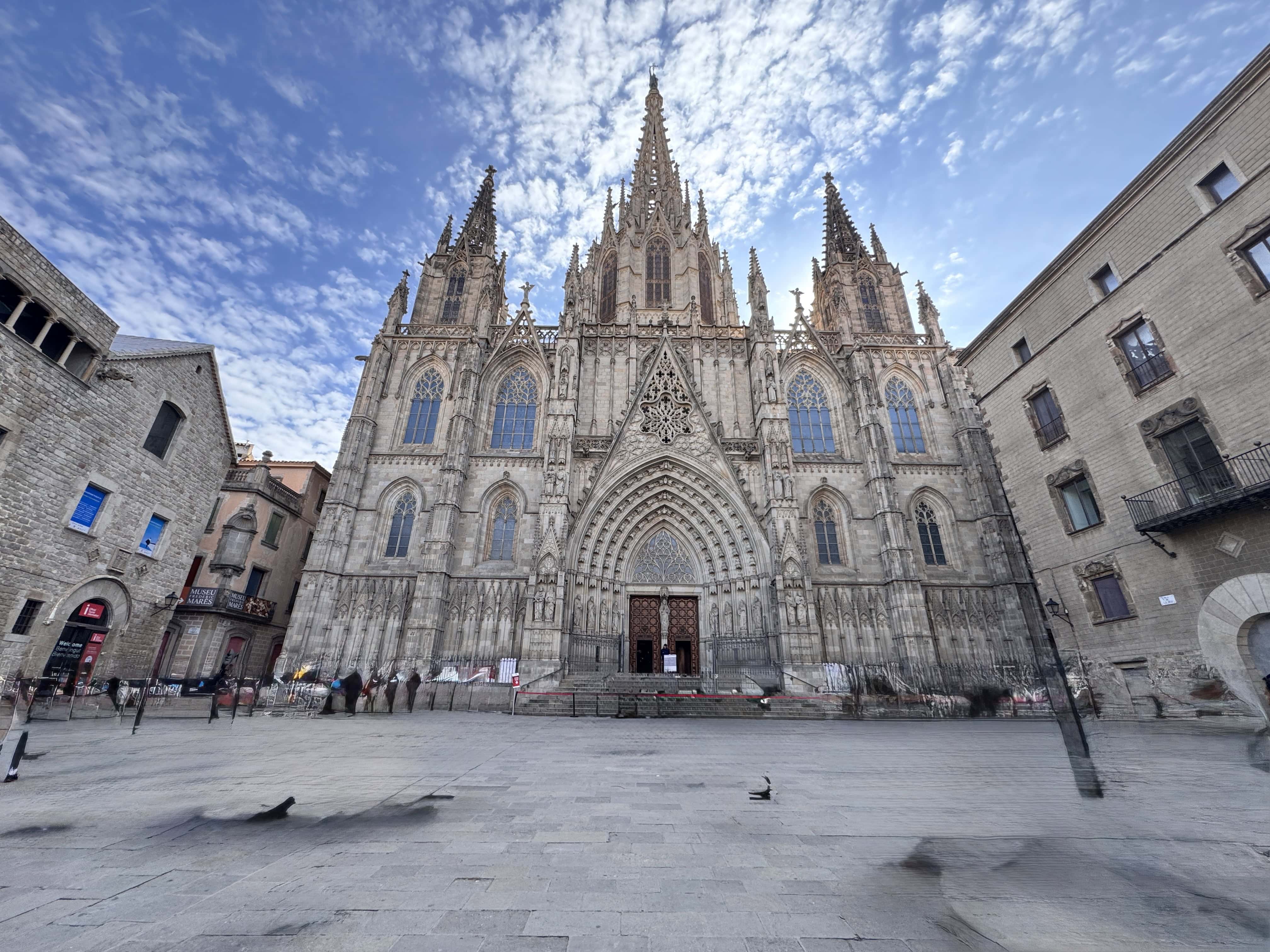 A large Gothic cathedral with ornate spires and detailed stonework stands in a spacious plaza under a partly cloudy sky, perfect for exploring on a Barcelona Family Vacation. Nearby, historic stone buildings frame the square where a few pigeons stroll.