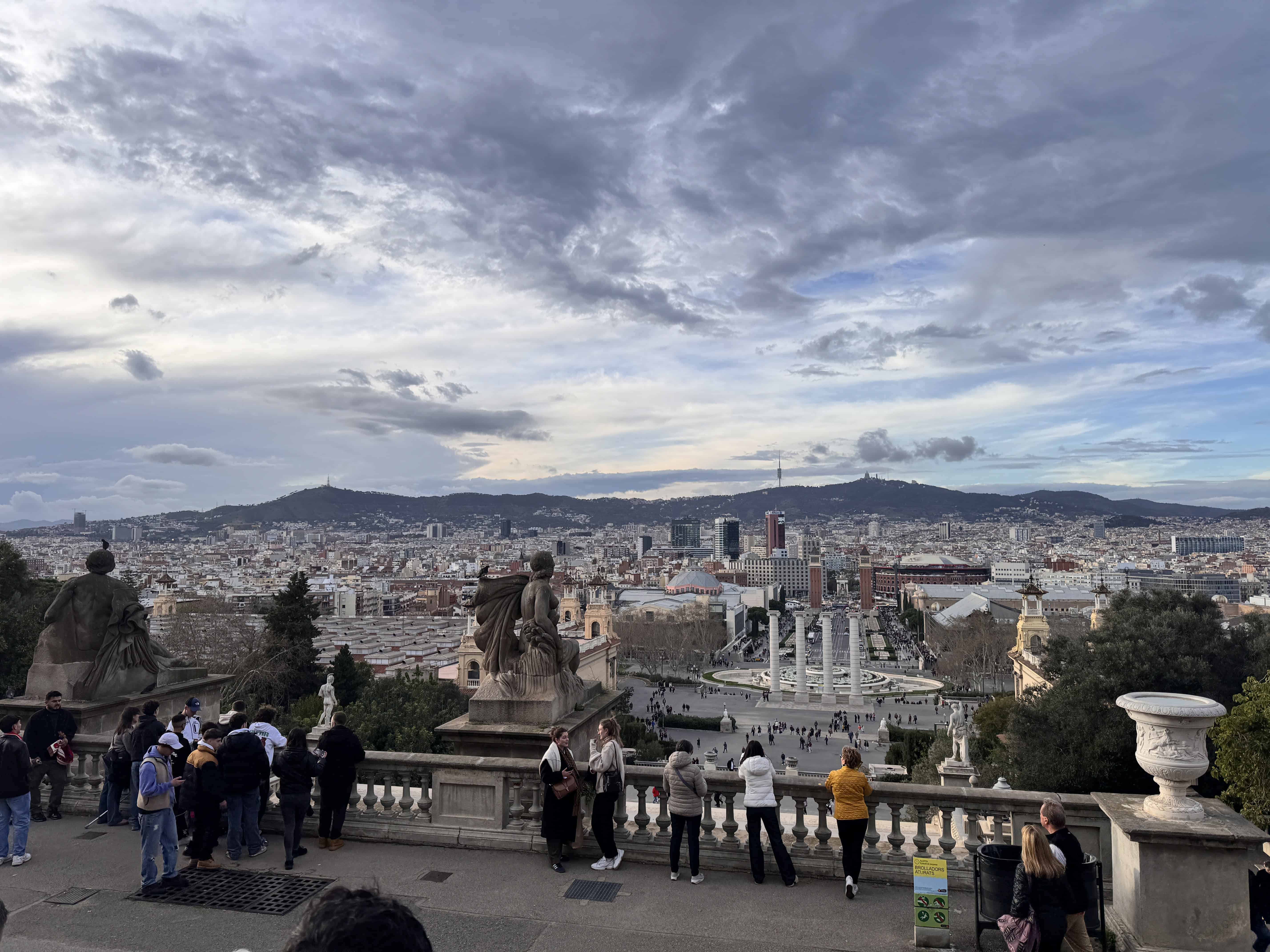 A family-friendly cityscape view of Barcelona from a high vantage point, with people standing on a terrace in the foreground, statues on either side, and buildings, hills, and a cloudy sky in the background.