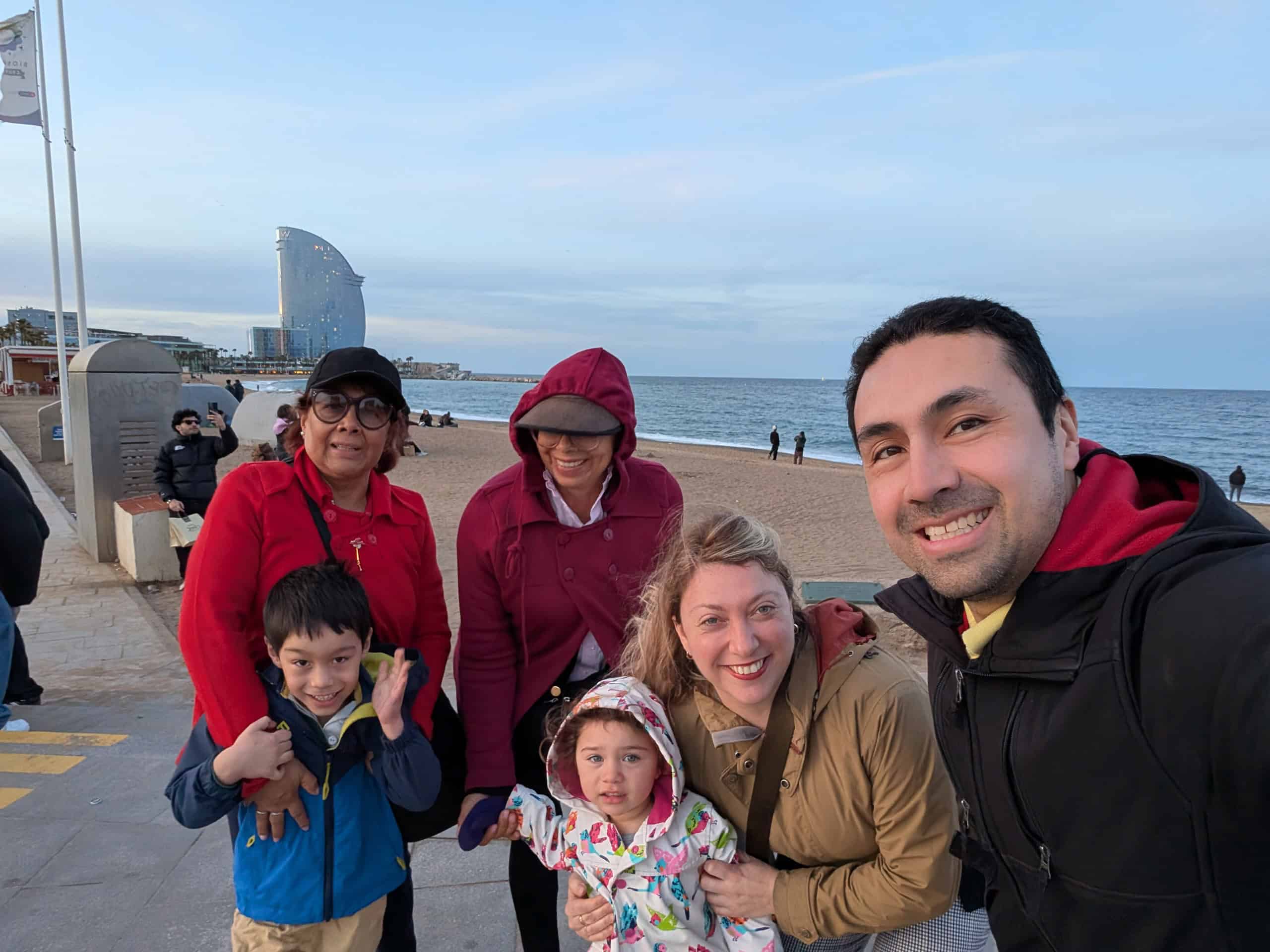 A group of six people, including two children, pose and smile for a photo on a beach promenade—capturing the joy of a Barcelona family vacation with the sea and a modern sail-shaped building in the background.