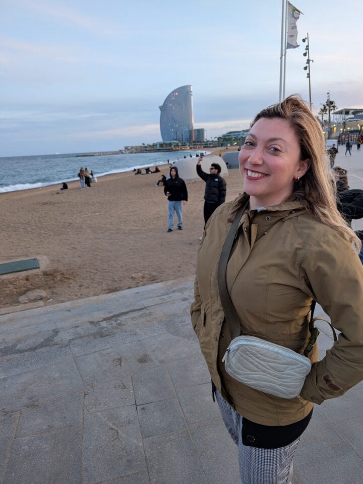 A woman in a tan jacket smiles at the camera while standing on a paved walkway by a beach—perfect for exploring Barcelona with kids. People enjoy the sand, and a modern glass building rises near the family-friendly shoreline.