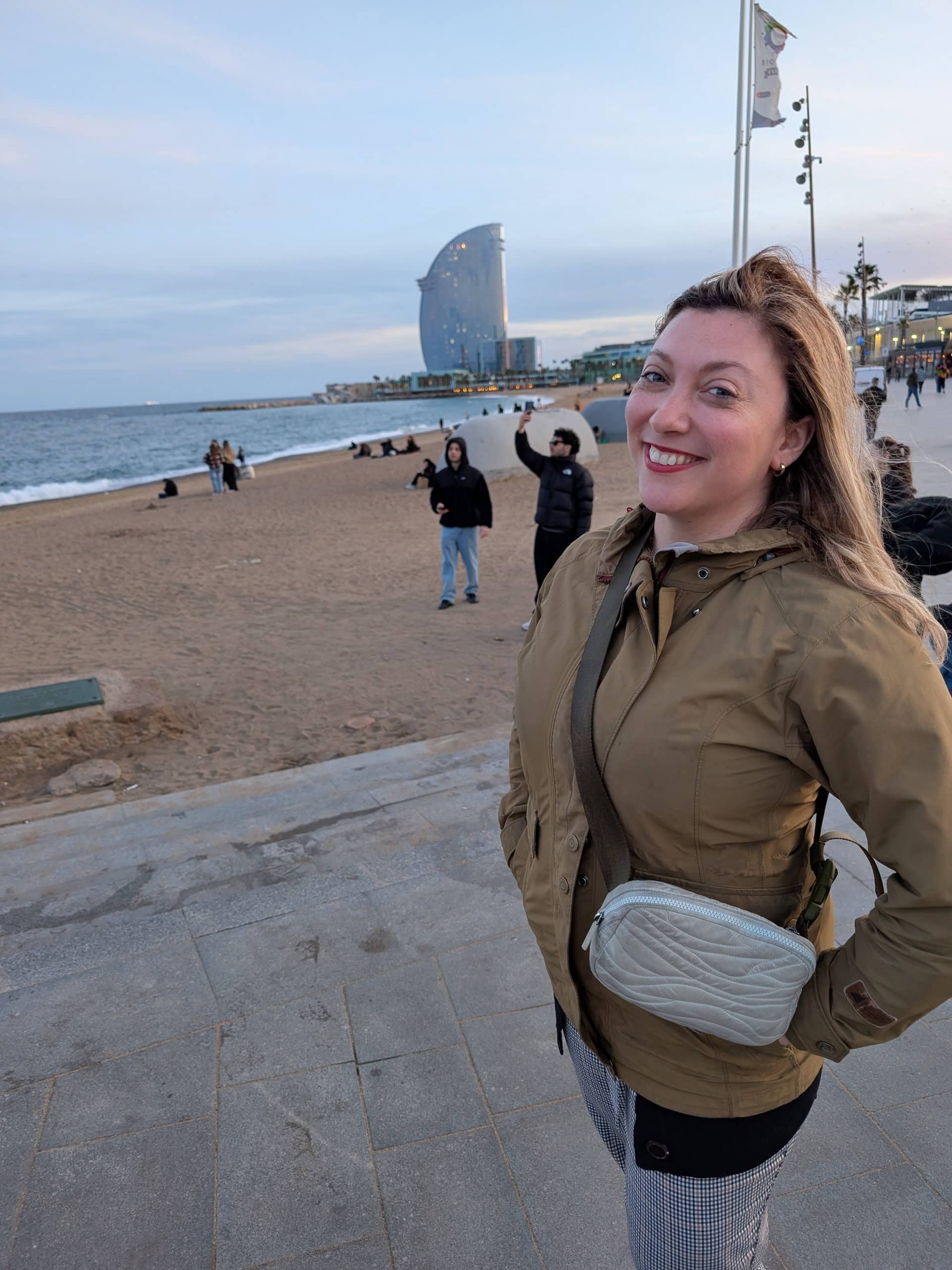 A woman in a tan jacket smiles at the camera while standing on a paved walkway by a beach—perfect for exploring Barcelona with kids. People enjoy the sand, and a modern glass building rises near the family-friendly shoreline.