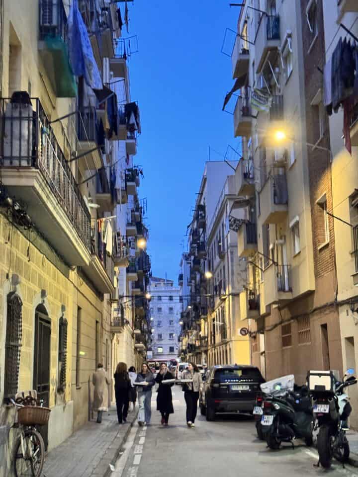 A narrow, family-friendly Barcelona street lined with tall apartment buildings, lit by streetlights at dusk. Several people walk in the middle of the street, while parked cars and motorcycles line both sides. Laundry hangs from some balconies.