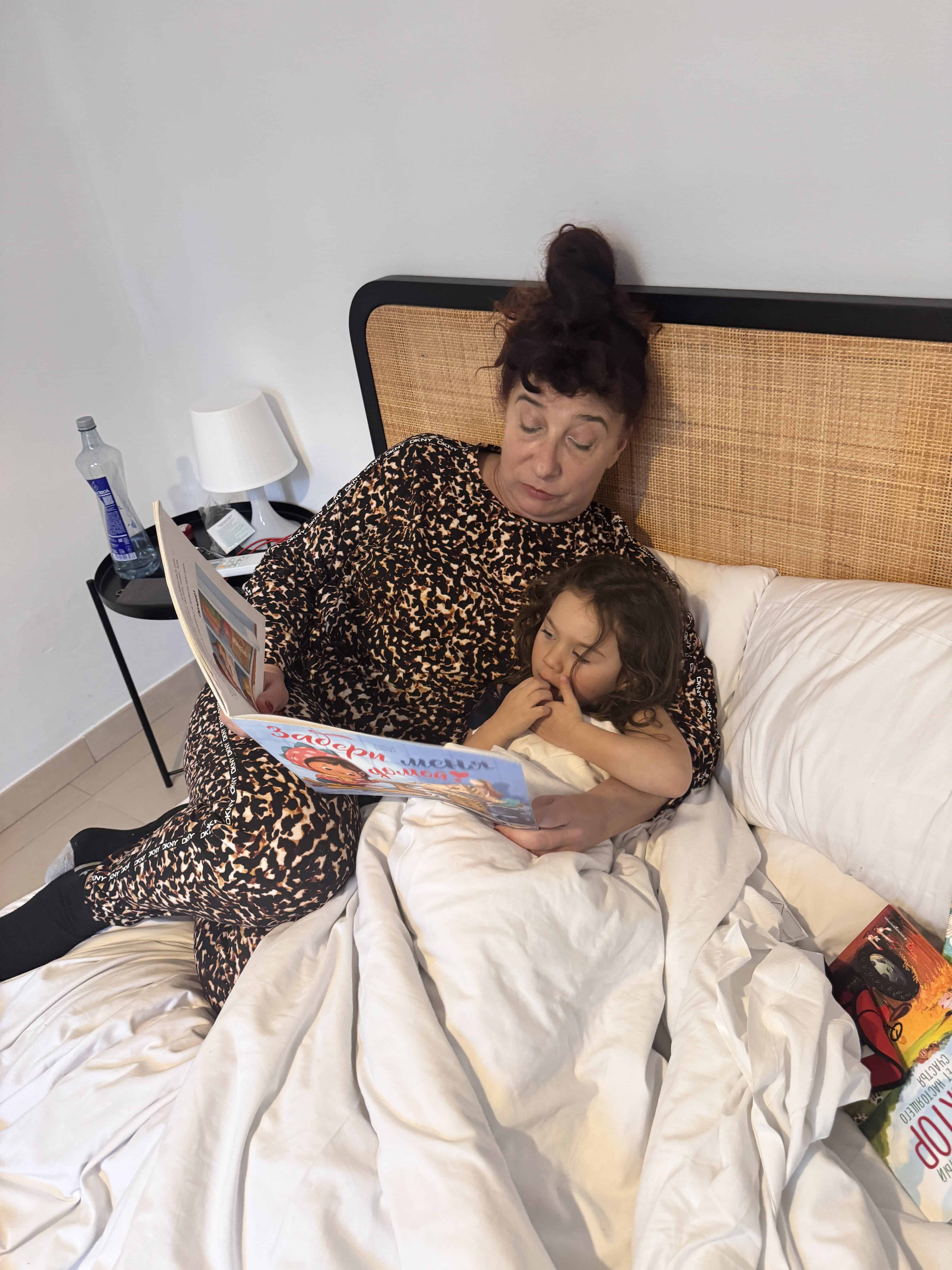 An adult in a leopard-print outfit sits in bed reading a picture book to a child, both snug under a white blanket. A lamp, water bottle, and snacks on the nightstand set the scene for this cozy Barcelona with Kids family moment.