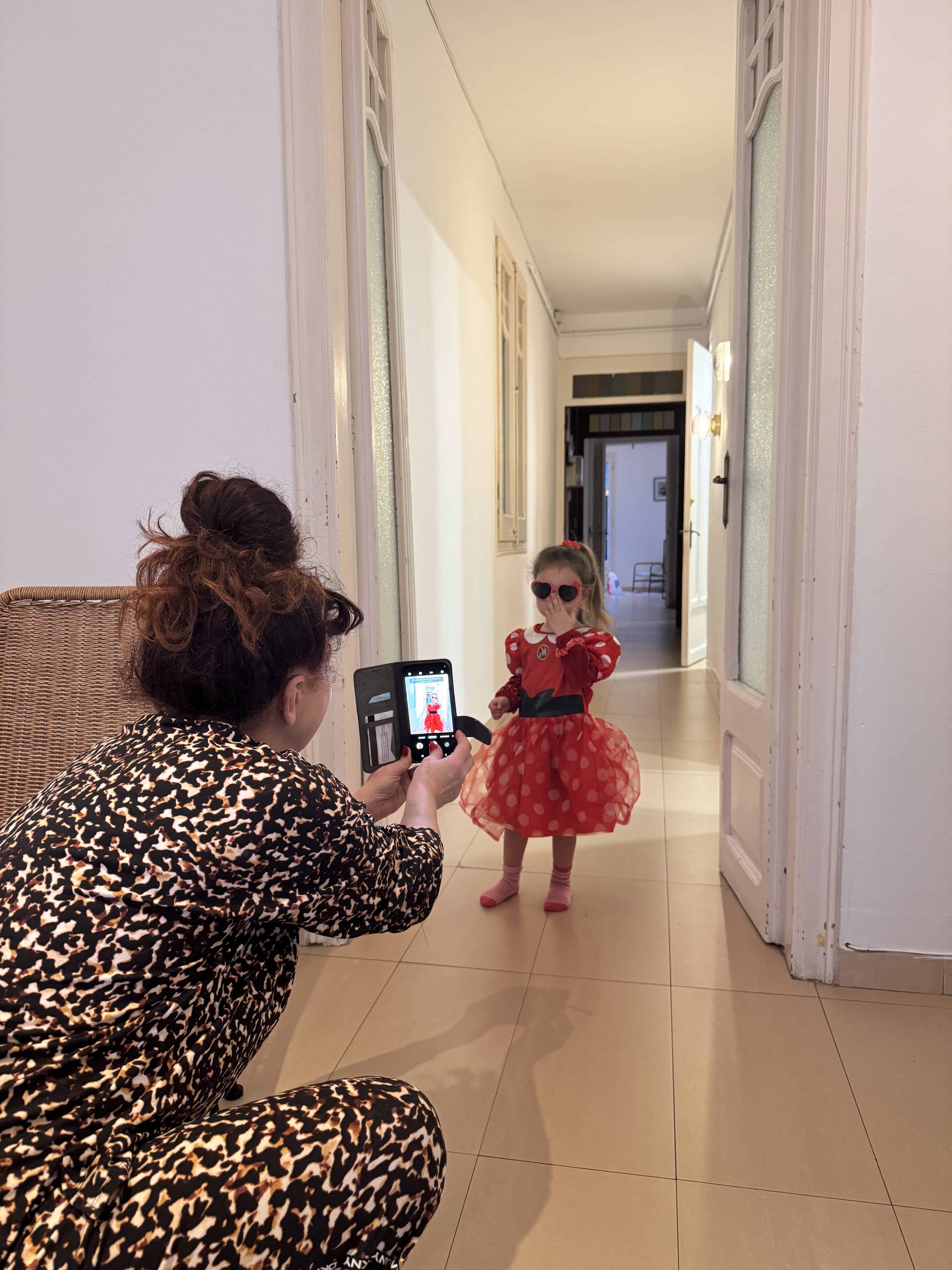 An adult kneels in a hallway taking a photo of a young child wearing a red dress, sunglasses, and holding a small handbag—capturing a stylish moment on their Barcelona Family Vacation in a well-lit indoor space.