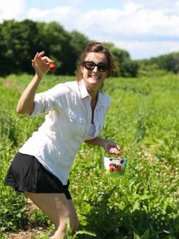 A woman wearing sunglasses, a white shirt, and a black skirt is picking strawberries in a green field, holding a small white bucket with strawberries and smiling at the camera.