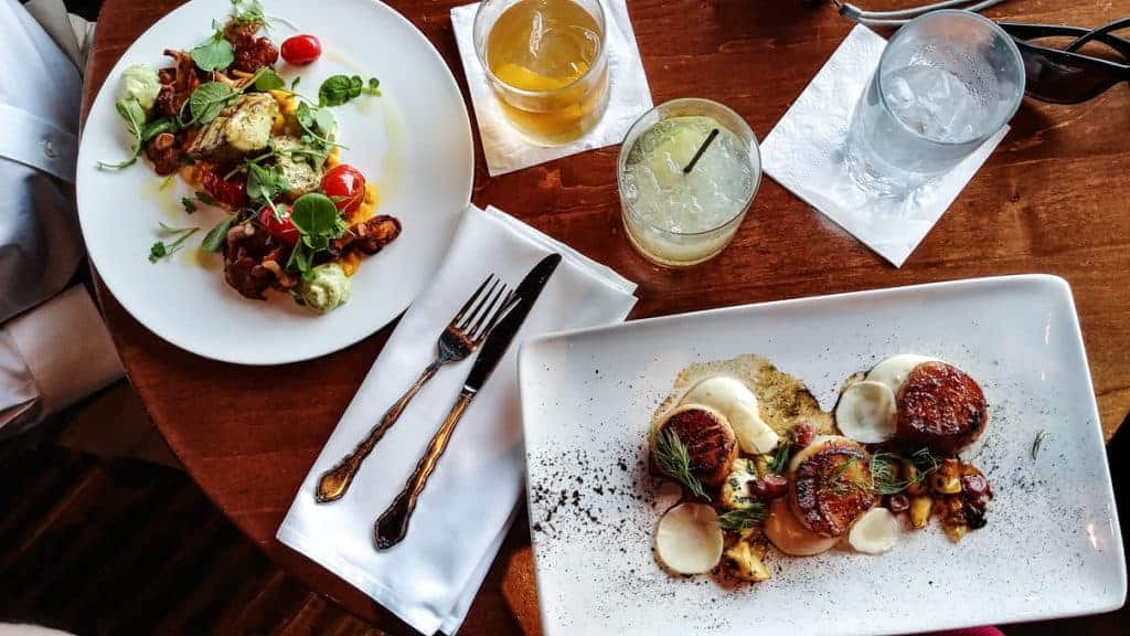 A wooden table with two plates of gourmet food, two cocktails, a glass of water, and utensils on napkins. One plate has seared scallops with garnish; the other features a mixed salad with tomatoes and greens.