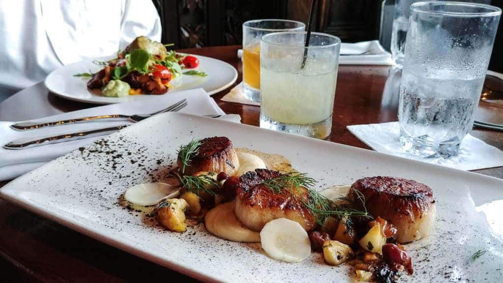 A rectangular white plate with three seared scallops on a bed of puree, garnished with herbs and diced vegetables. In the background are a salad, a cocktail, and glasses of water on a wooden table.