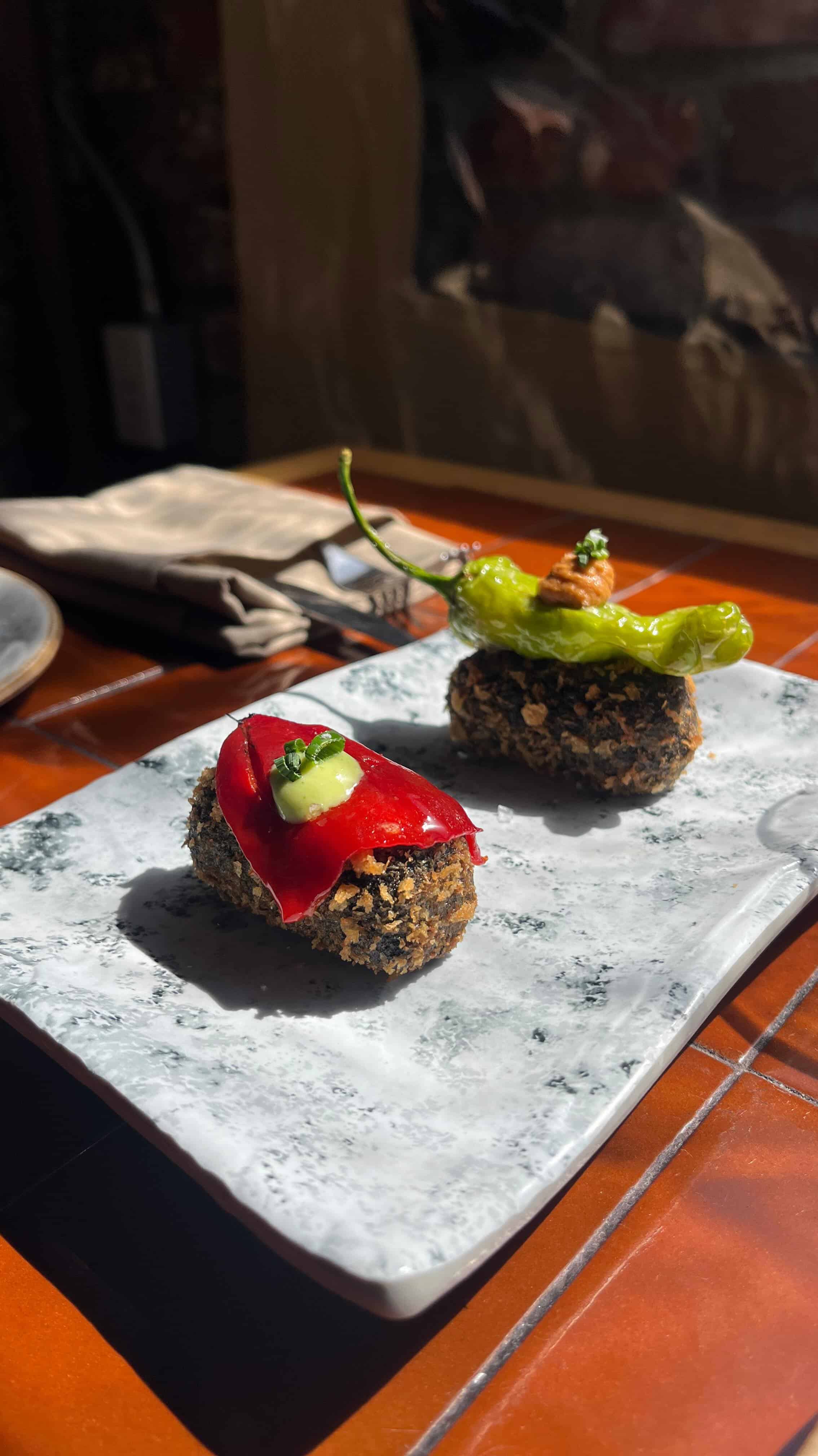 Two gourmet appetizers on a rectangular marble plate, each with a breaded base and vibrant garnishes&mdash;one featuring red pepper, another green&mdash;perfect for savoring during your 4 days in Barcelona on a sunlit table.