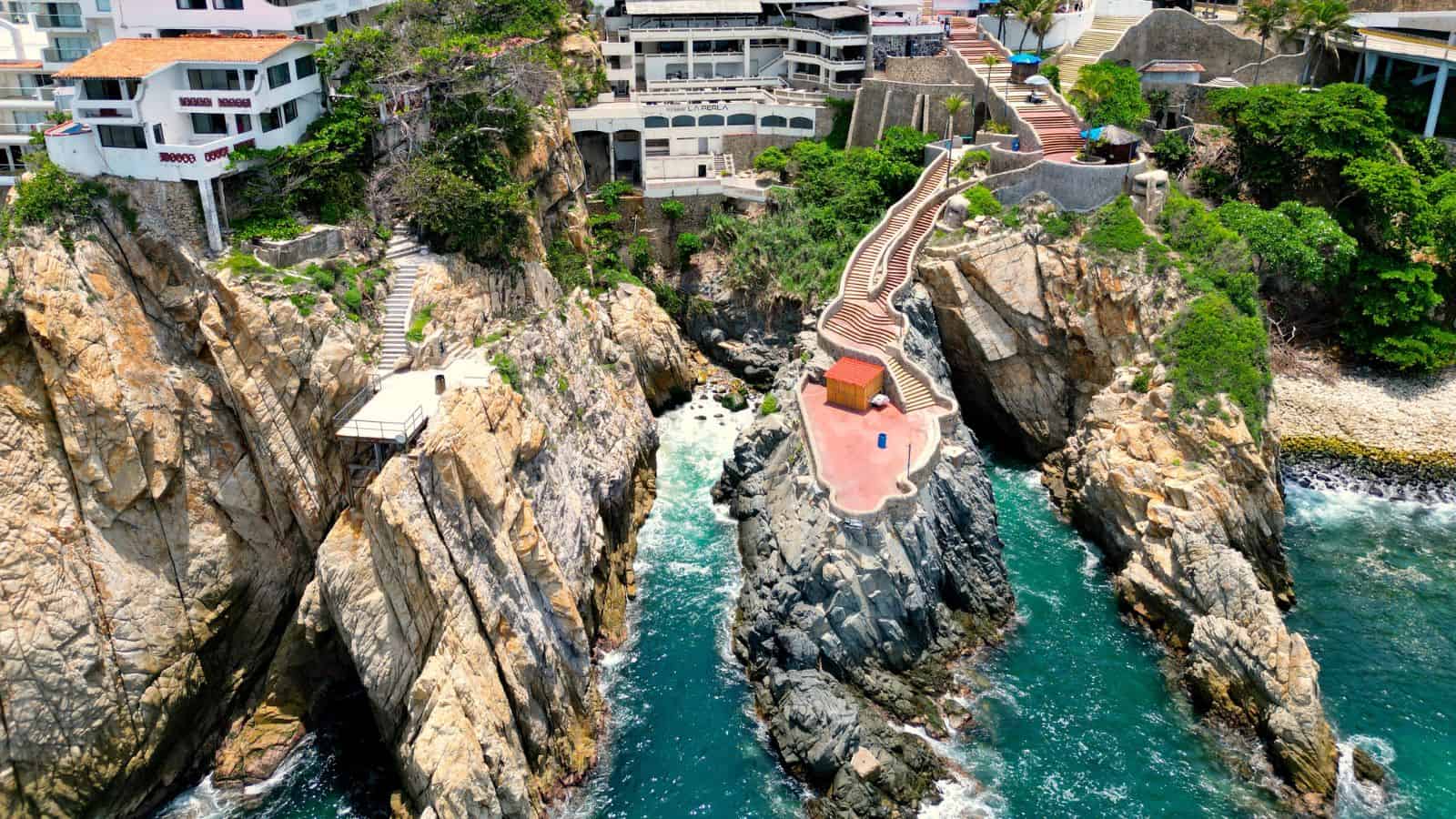 Aerial view of a rocky coastline with a platform and staircase extending over the rocks toward the ocean, surrounded by buildings and lush greenery in a coastal urban area.