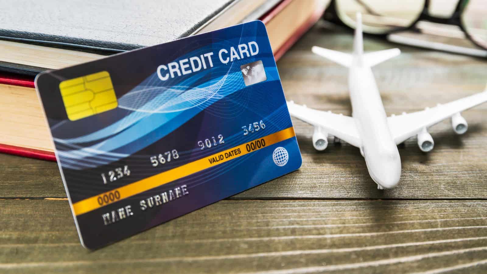 A credit card stands upright in front of a closed book and a small model airplane on a wooden surface, with part of a pair of eyeglasses visible in the background.