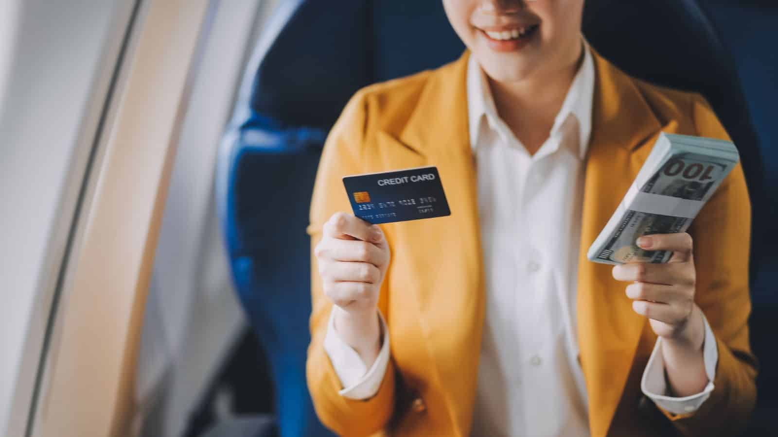 A person wearing a yellow blazer sits on an airplane seat, smiling while holding a credit card in one hand and several $100 bills in the other.