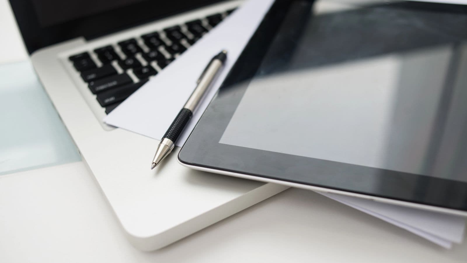 A close-up view of a laptop keyboard, a tablet, a silver and black pen, and sheets of paper overlapping on a white desk.