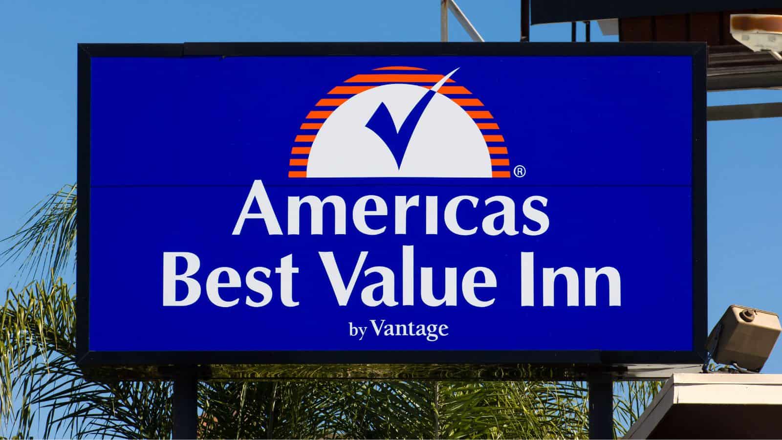 A blue sign with a white and orange logo displays the words "Americas Best Value Inn by Vantage" in white capital letters—a name sometimes mentioned among US hotel chains disappointing guests. Palm tree leaves and a security camera are visible in the background.