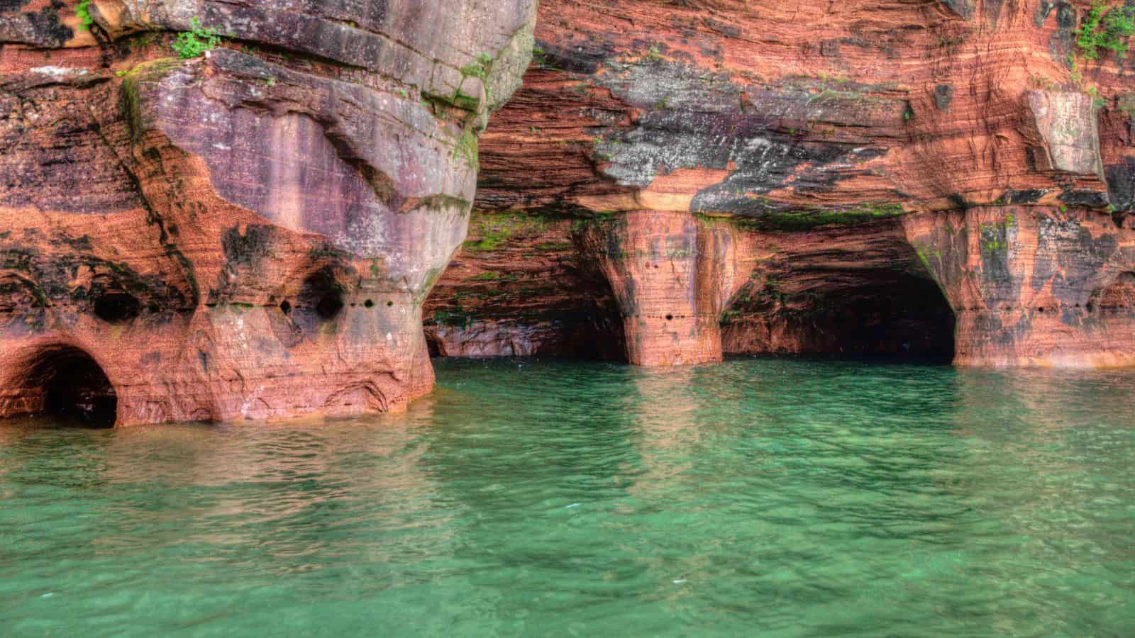 A view of reddish-brown rocky cliffs with arch-shaped sea caves and openings at the waterline, above clear green water. Moss and weathering are visible on the rock surfaces.