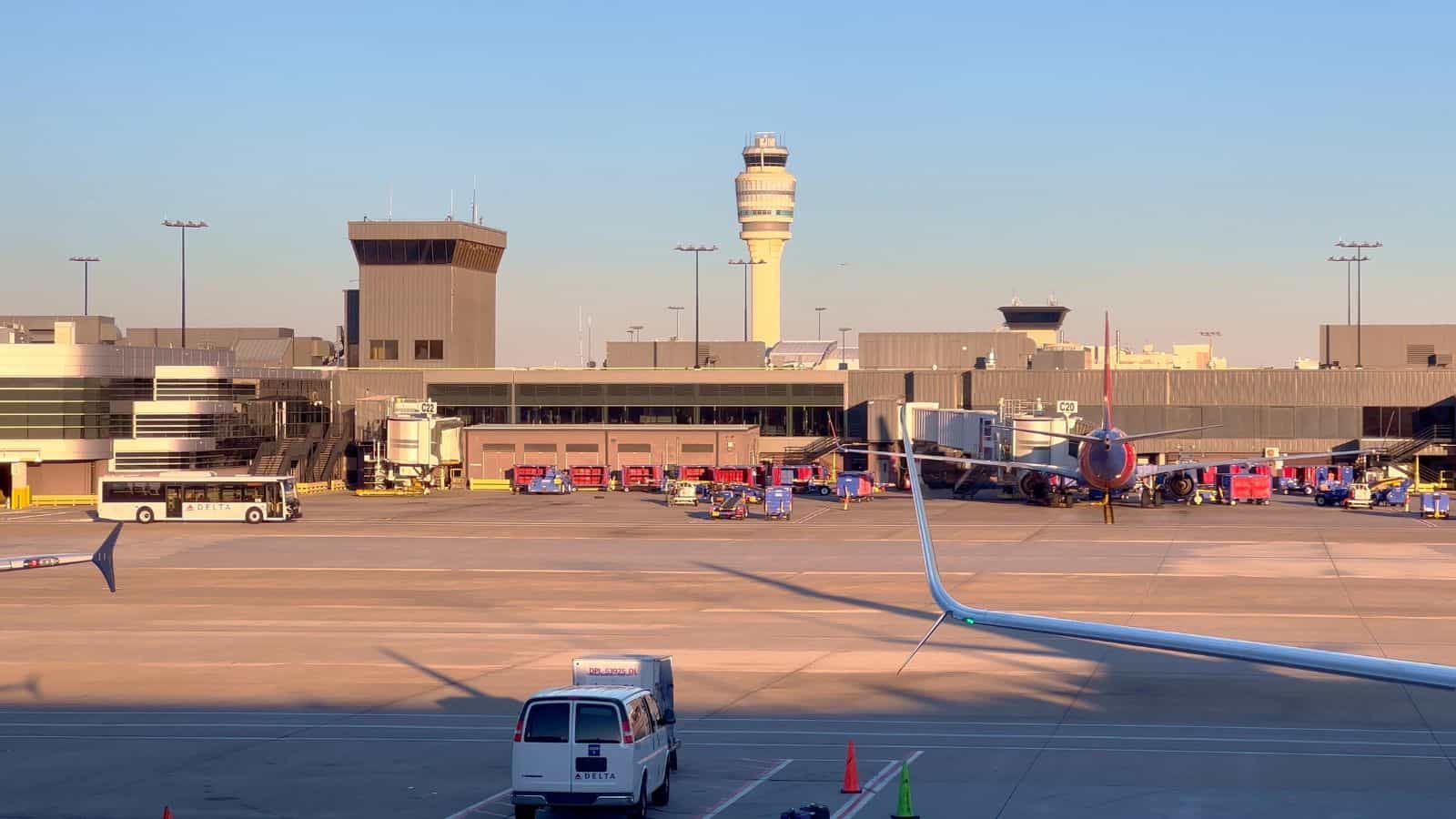 Airport tarmac with parked airplanes near terminal gates, ground vehicles, and airport staff visible. Control tower and airport buildings are in the background under a clear sky.