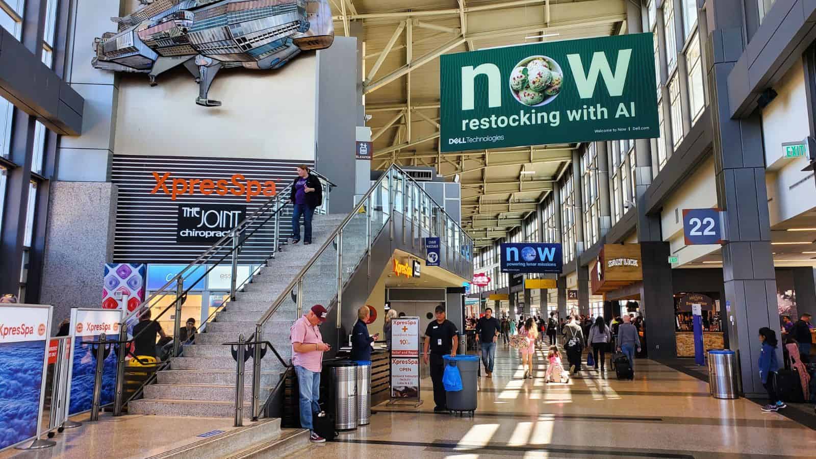 A busy airport terminal with people walking and standing, suitcases nearby, large signs overhead advertising AI restocking, and storefronts including a spa and restaurants. Natural light enters through the high glass ceiling.