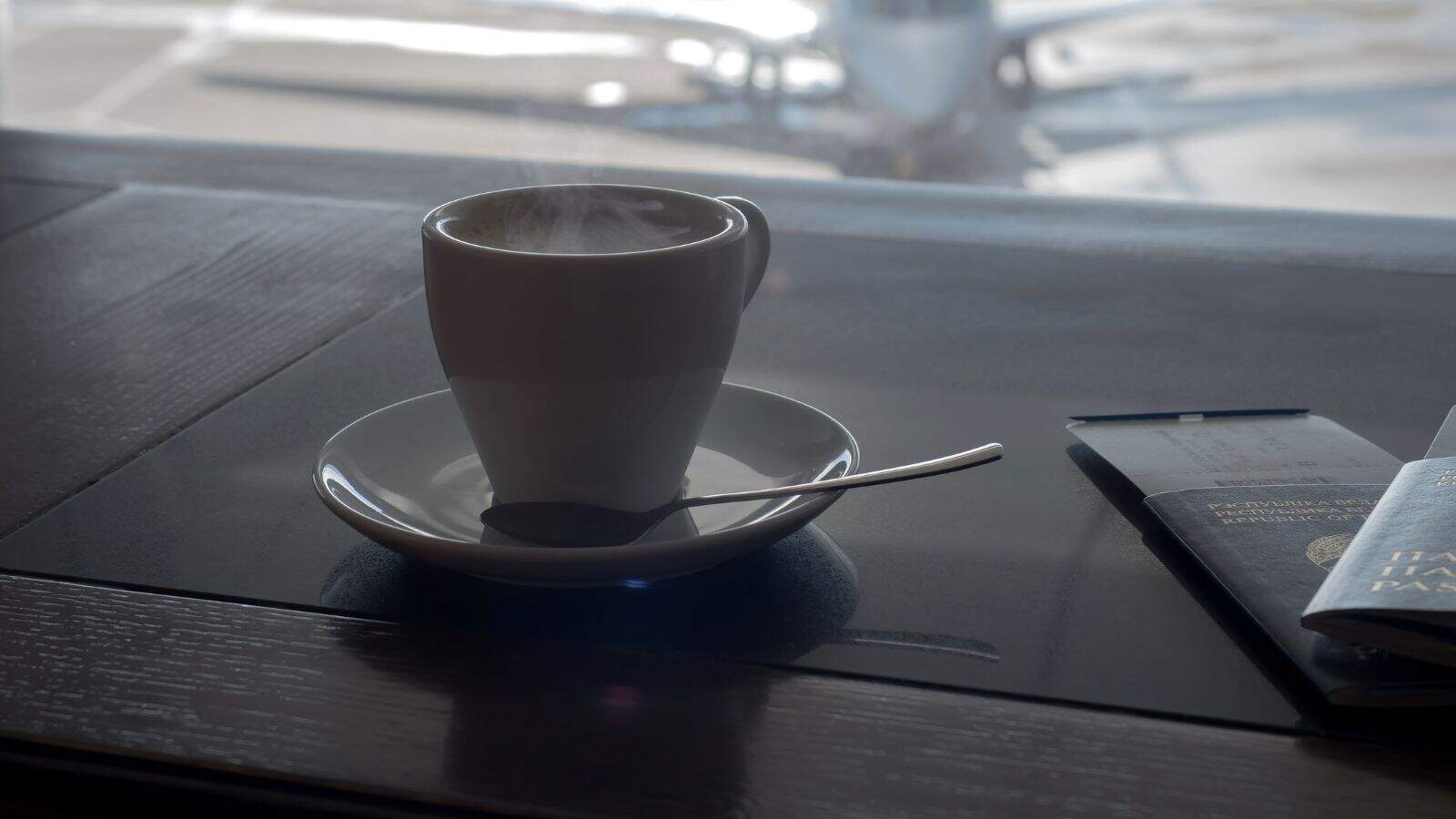 A steaming cup of coffee sits on a white saucer with a spoon as a closed passport and boarding pass rest nearby, suggesting someone preparing to conquer their fear of flying while an airplane waits outside the window.
