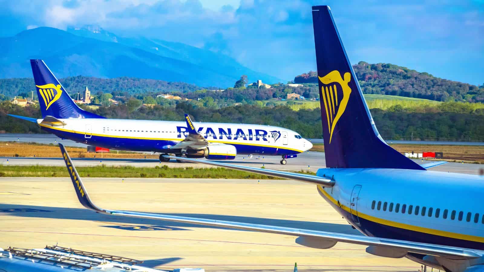 A Ryanair airplane taxis on a runway at an airport, with another Ryanair aircraft in the foreground and green hills with buildings in the background under a partly cloudy sky.