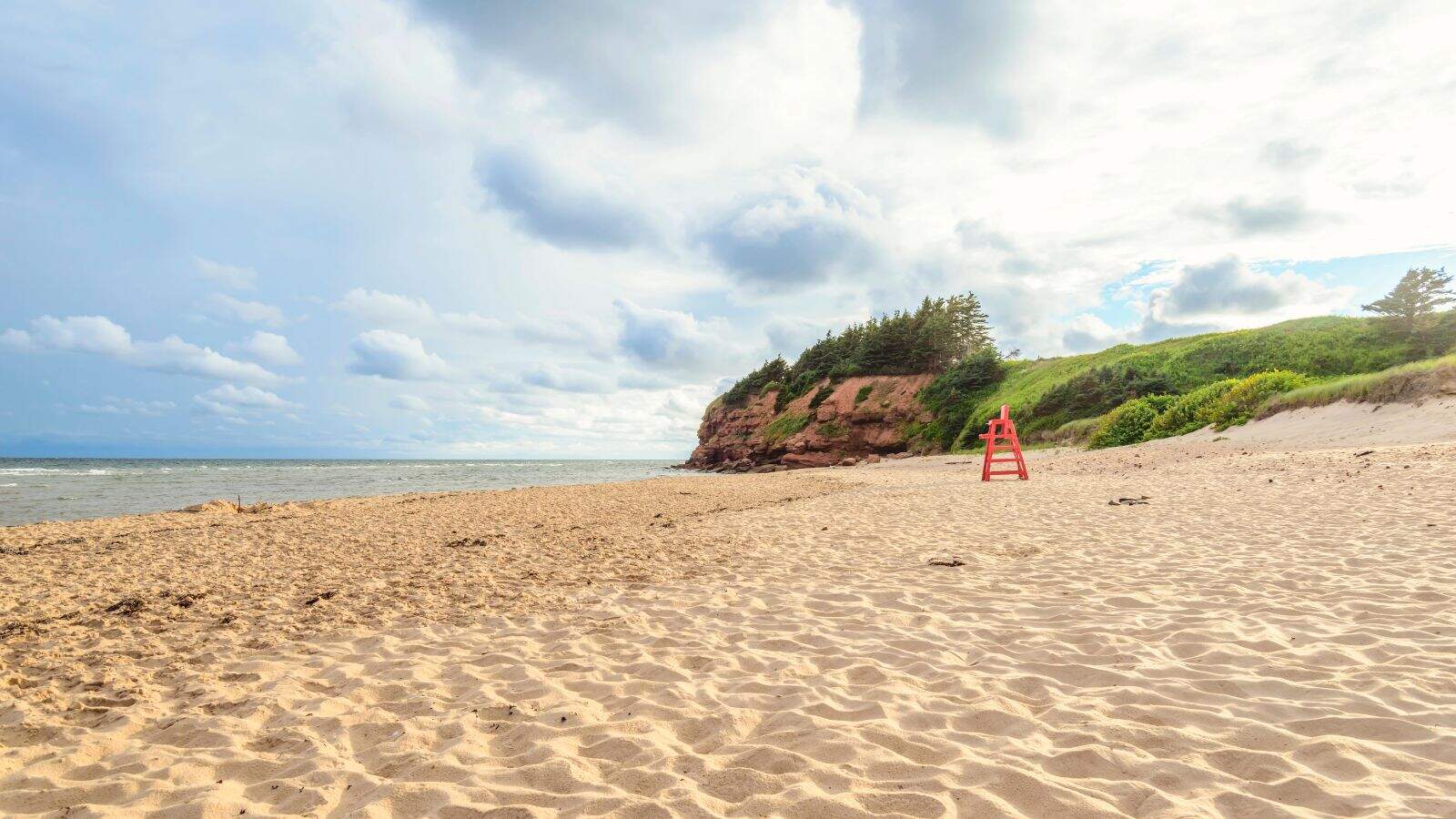 A sandy beach with scattered footprints, a red lifeguard chair, and grassy cliffs in the background under a partly cloudy sky. The ocean is visible on the left, and greenery covers the cliff.