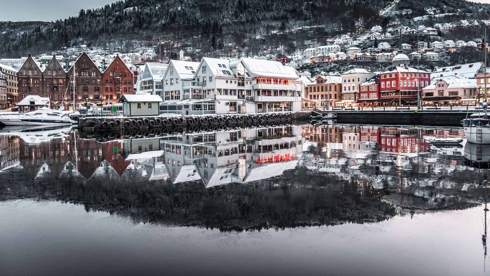 Colorful wooden buildings and boats line a snowy waterfront, their reflections visible in the calm water. A hillside covered with snow and homes rises in the background under an overcast sky.