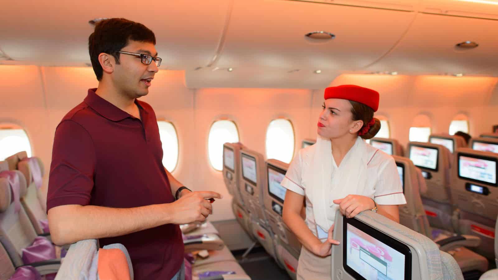A man in a maroon polo shirt speaks to a flight attendant in a white uniform and red hat inside an airplane cabin. The scene captures methods to conquer fear of flying, with comforting staff, personal screens, and soft lighting.