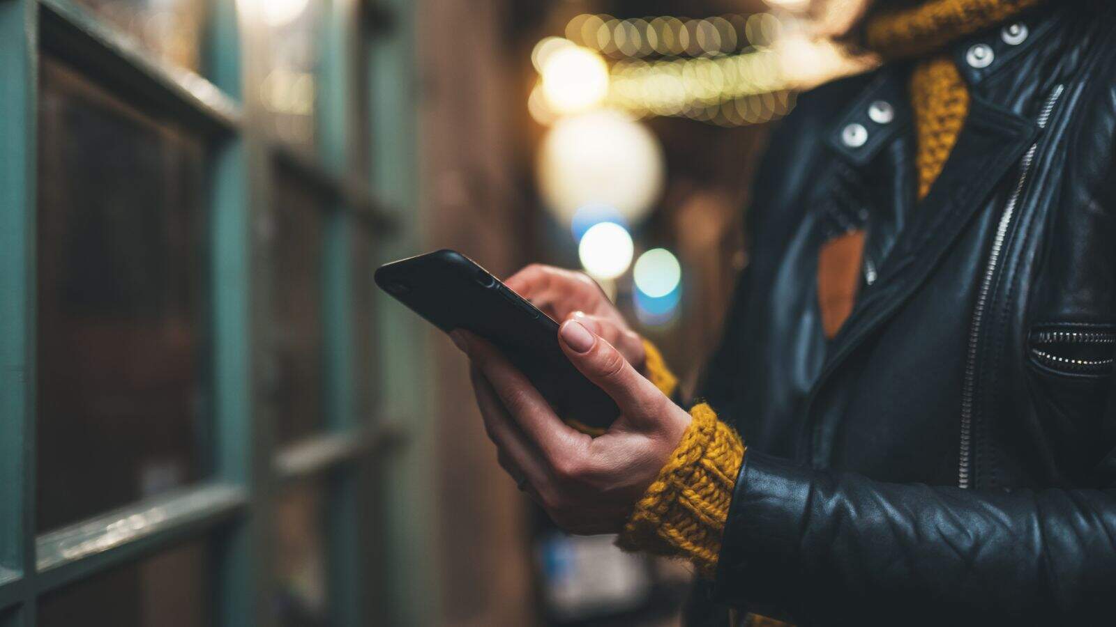 A person wearing a black leather jacket and yellow knit sweater is holding and using a smartphone outside at night, with blurred lights in the background.