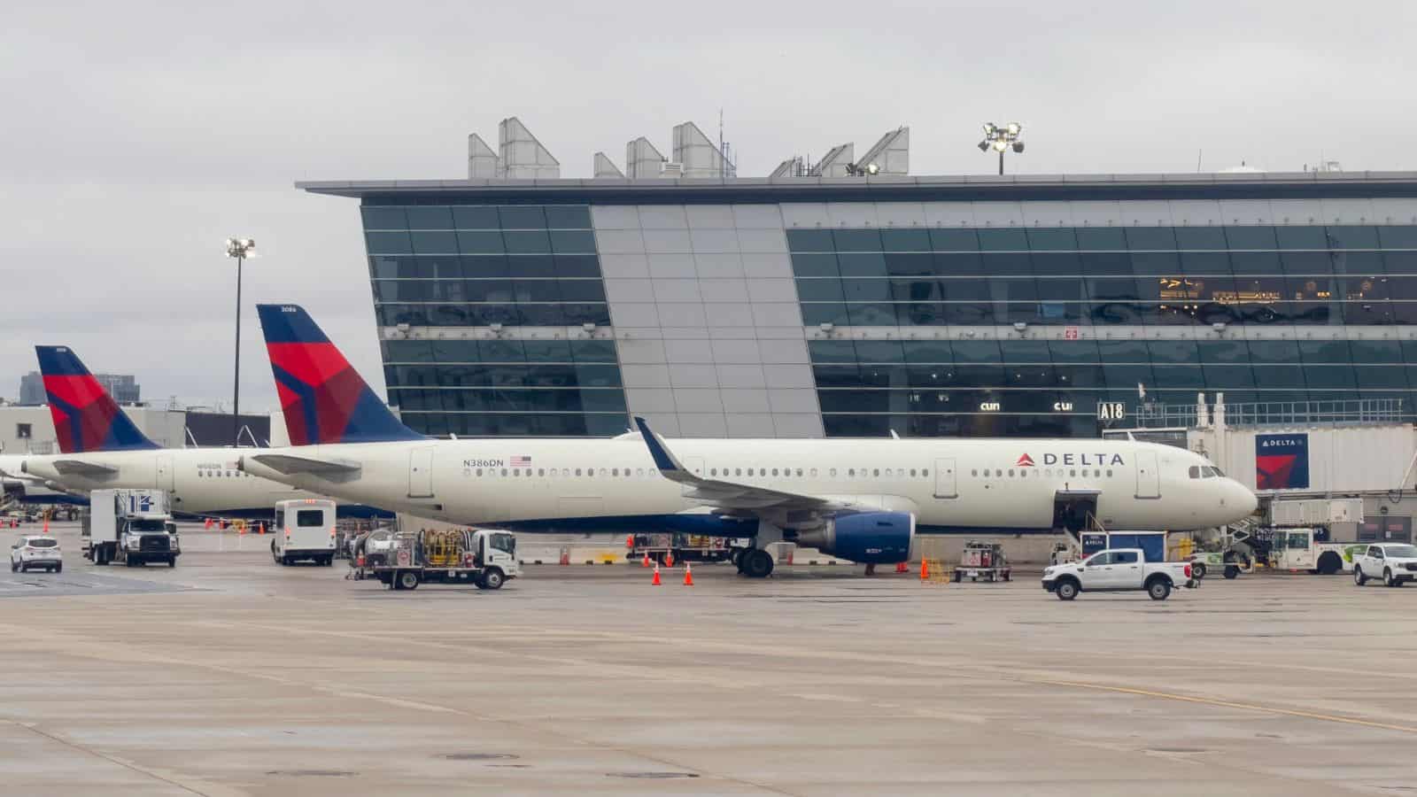 Two Delta Air Lines planes are parked at airport gates near a modern terminal building on a cloudy day. Ground vehicles and equipment are positioned around the planes on the wet tarmac.