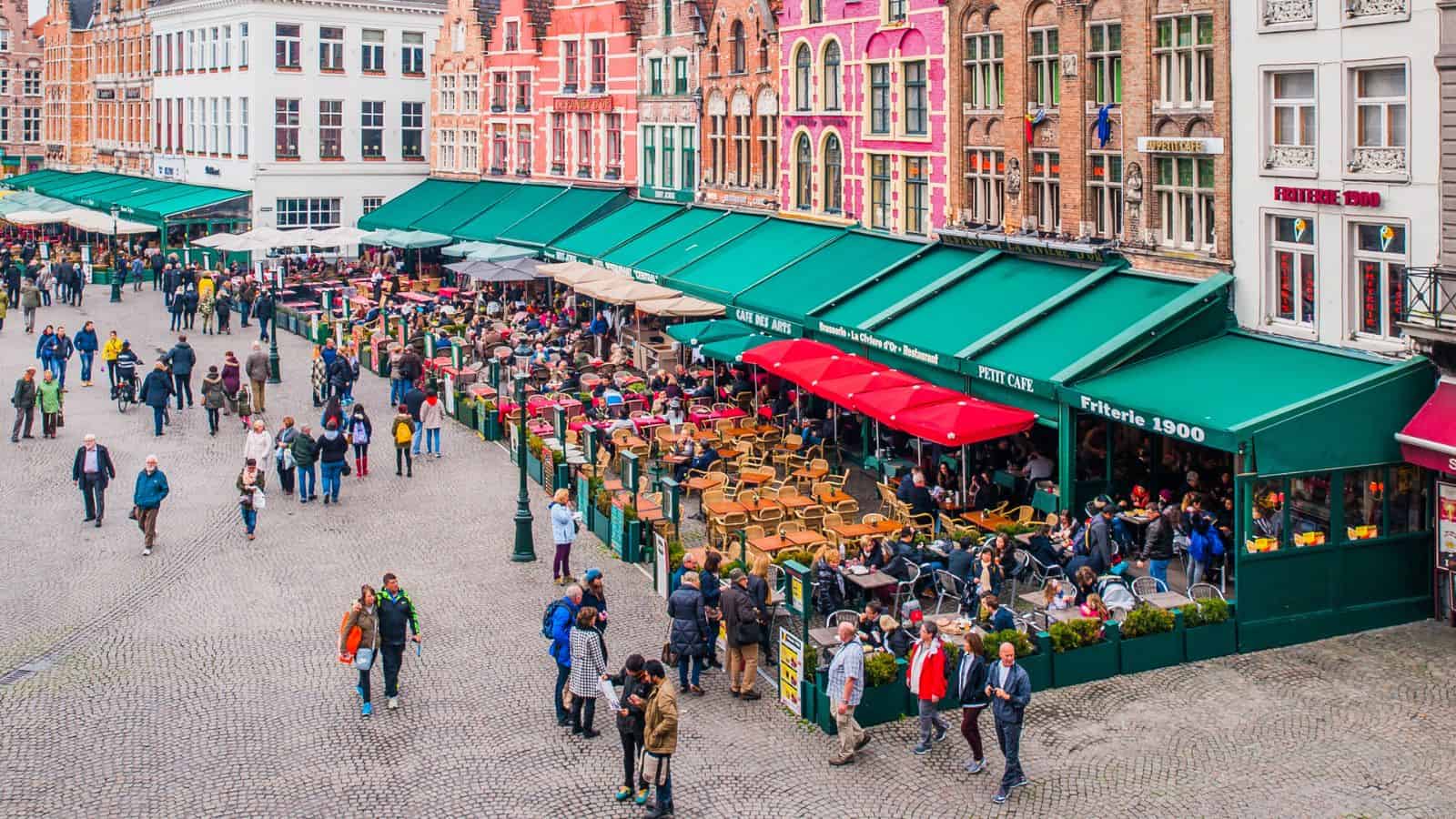 A busy outdoor café in a European square, with rows of colorful buildings and green awnings. People are seated at tables and walking along the cobblestone street, creating a lively atmosphere.