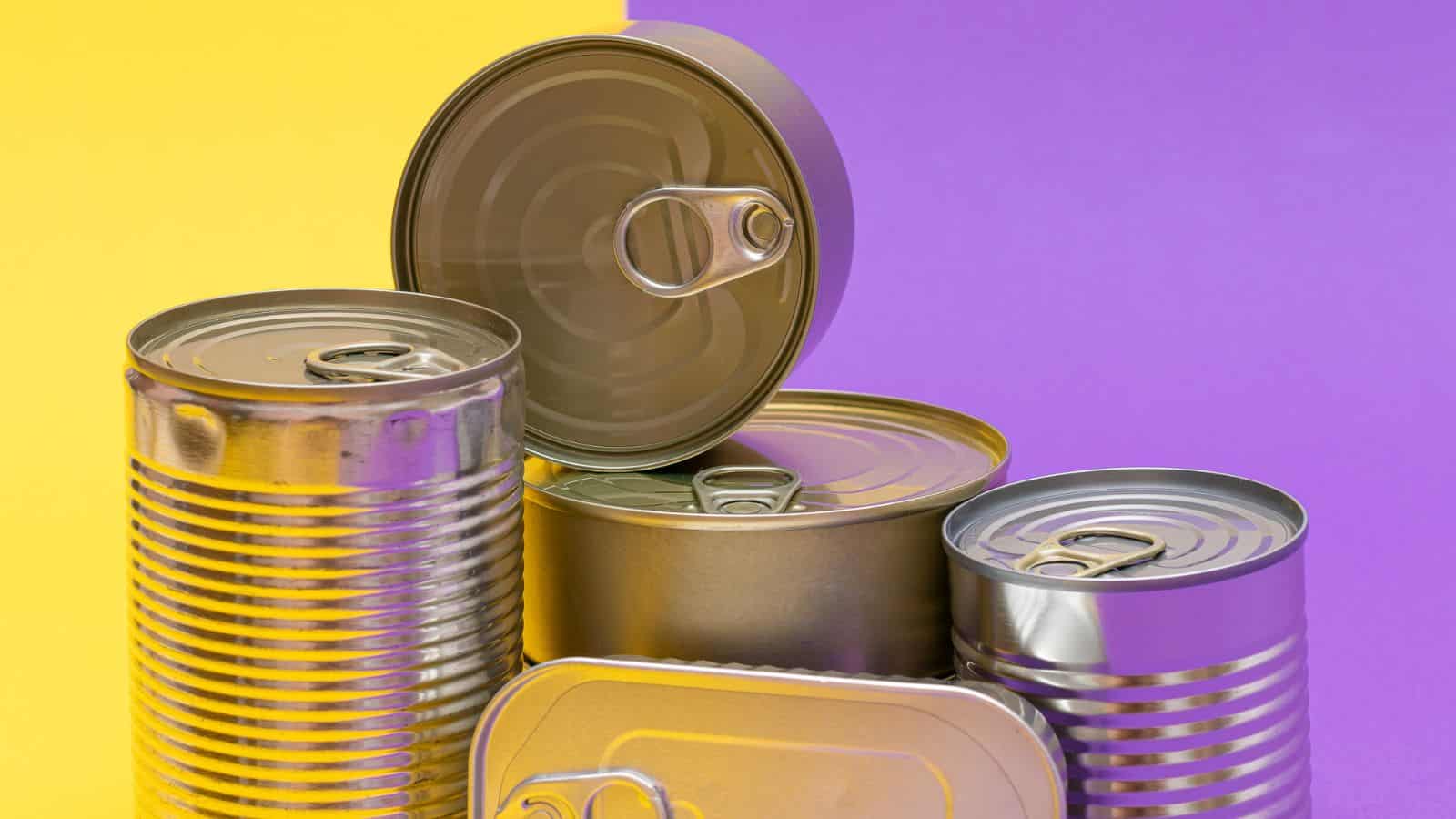 Several unopened metal cans with pull-tab lids are arranged in front of a split yellow and purple background. The cans vary in size and shape, and one can is placed on top with its lid facing upward.