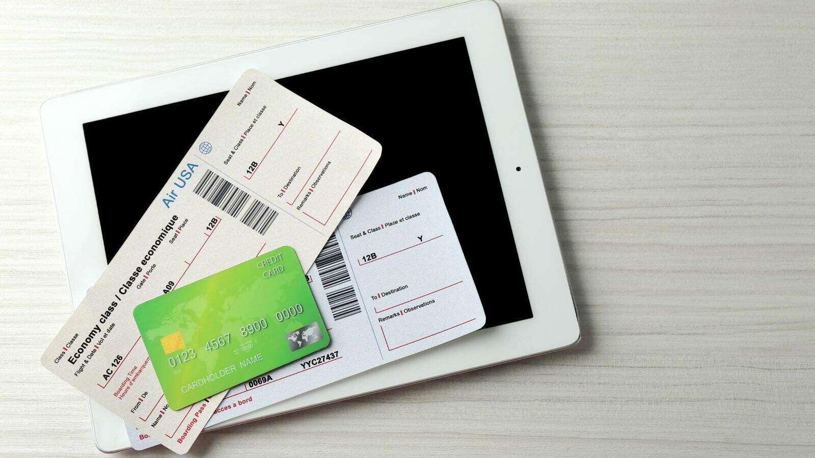 A tablet lies on a light-colored wooden surface with two airline boarding passes and a green credit card placed on top of it. The tablet screen is black and facing upward.