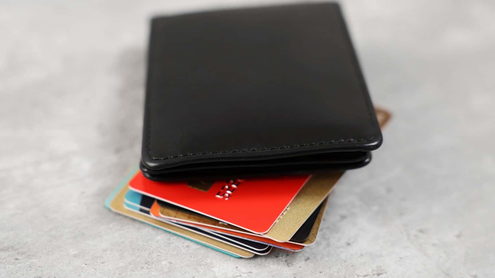 A black leather wallet is partially open on a gray surface, revealing several colorful plastic cards, including red, gold, and patterned ones, sticking out from the wallet.