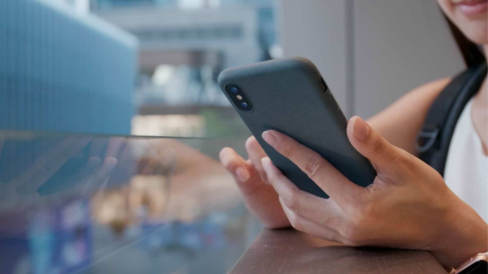 A person holding a smartphone with both hands while standing next to a glass railing indoors. The screen is not visible, and their face is mostly out of frame.