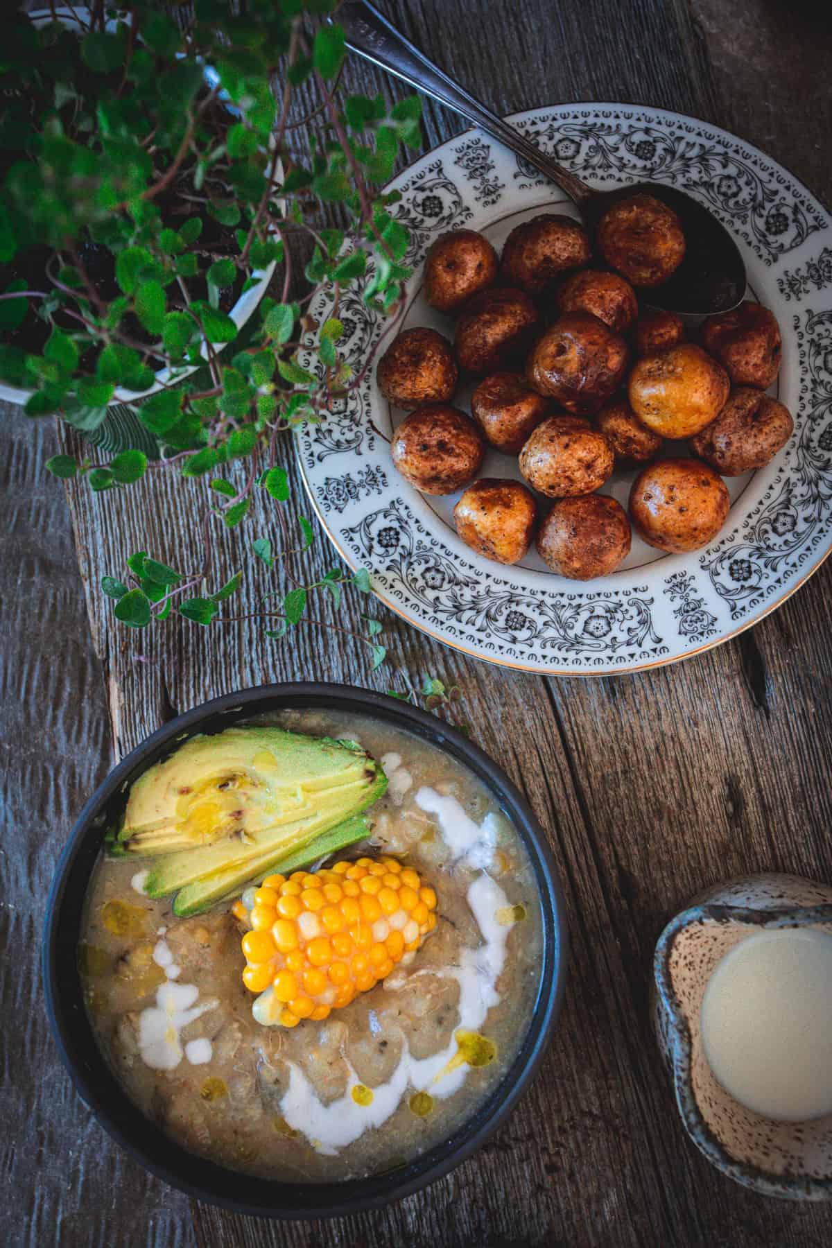 Bowl of soup with corn and avocado, plate of roasted potatoes, and glass of milk on a rustic table—classic elements of traditional vegetarian Colombian dishes.