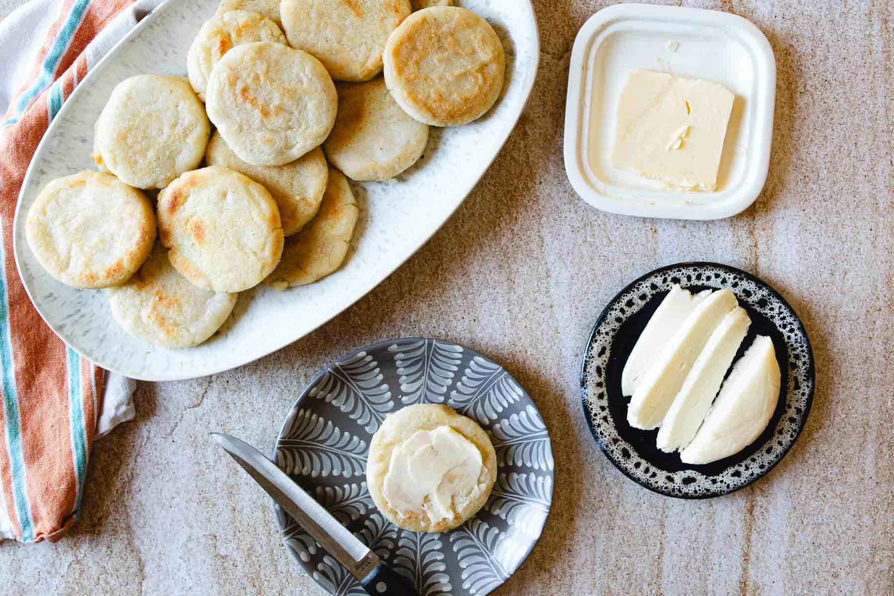 Plate of golden arepas, a pat of butter, a cheese wedge, and a buttered arepa on a small plate with a knife—classic elements of traditional vegetarian Colombian dishes.