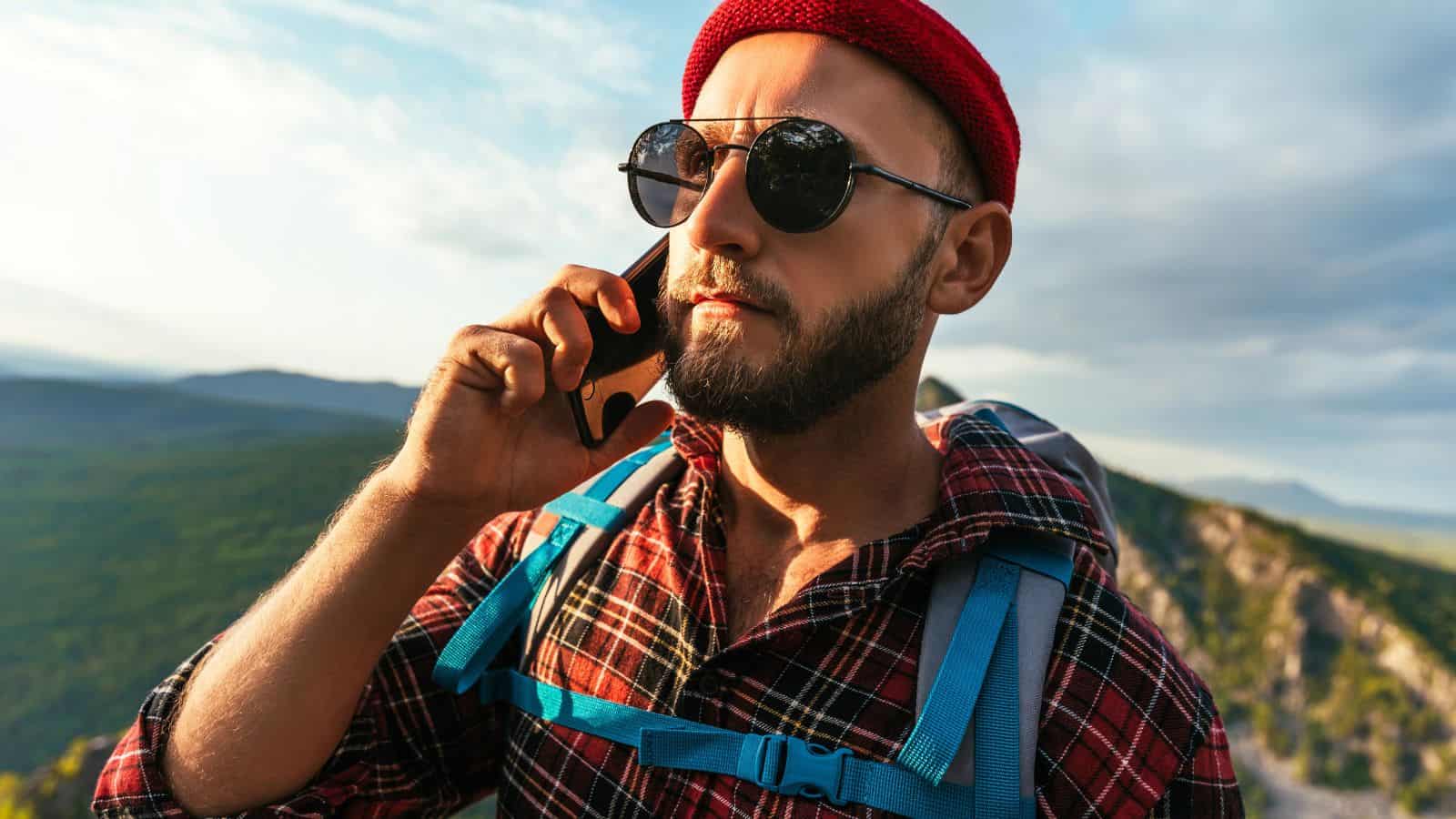 A man wearing sunglasses, a red beanie, and a plaid shirt with a blue backpack stands outdoors on a mountain trail, talking on a smartphone with a scenic landscape in the background.