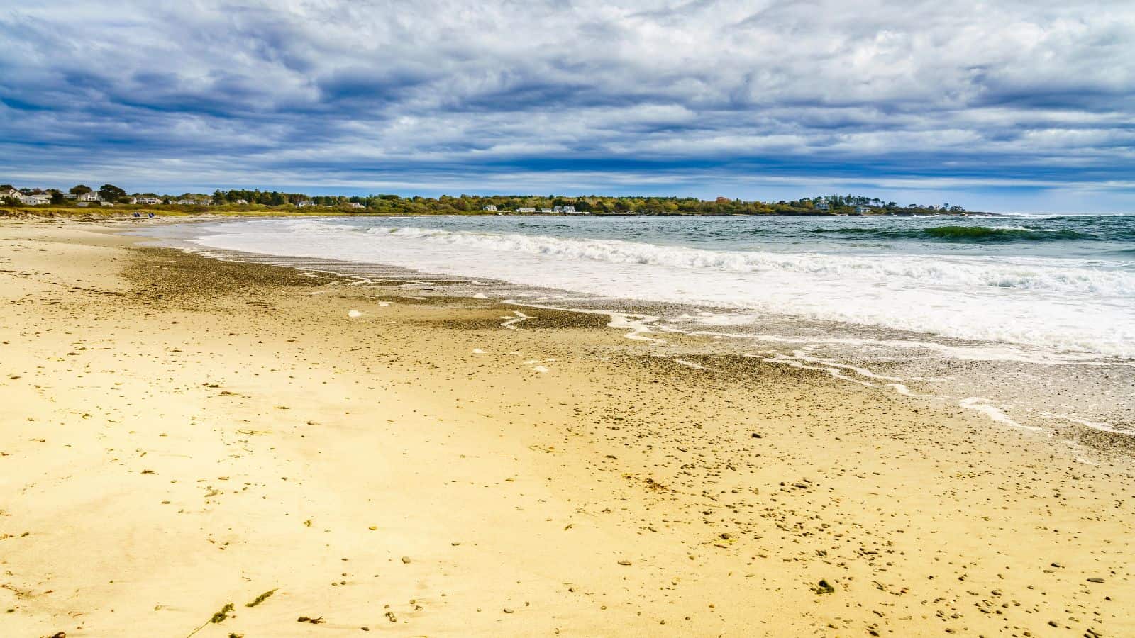 Wide sandy beach with gentle waves washing ashore under a mostly cloudy sky. Low, distant tree line and some buildings are visible along the horizon in the background.