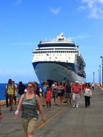 A large cruise ship is docked at a pier. Numerous people are walking along the pier, some in casual clothes, with the ocean and blue sky in the background.