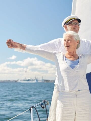 An older couple stands at the bow of a sailboat, arms outstretched and smiling. The man wears a captain’s hat. The water and city skyline are visible in the background on a sunny day.