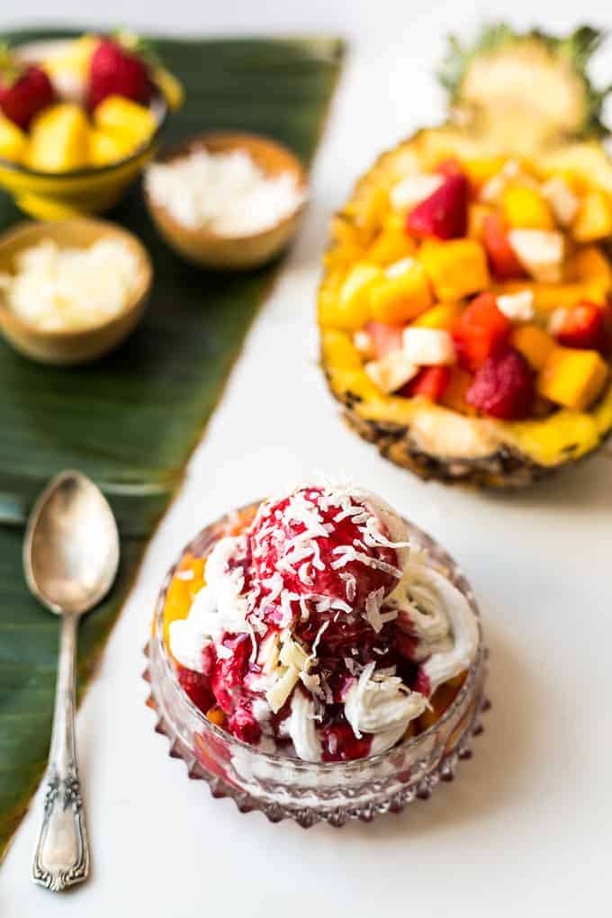 A tropical fruit sundae with coconut and berries in a glass bowl, reminiscent of traditional vegetarian Colombian dishes, with fresh fruit and a pineapple boat in the background.
