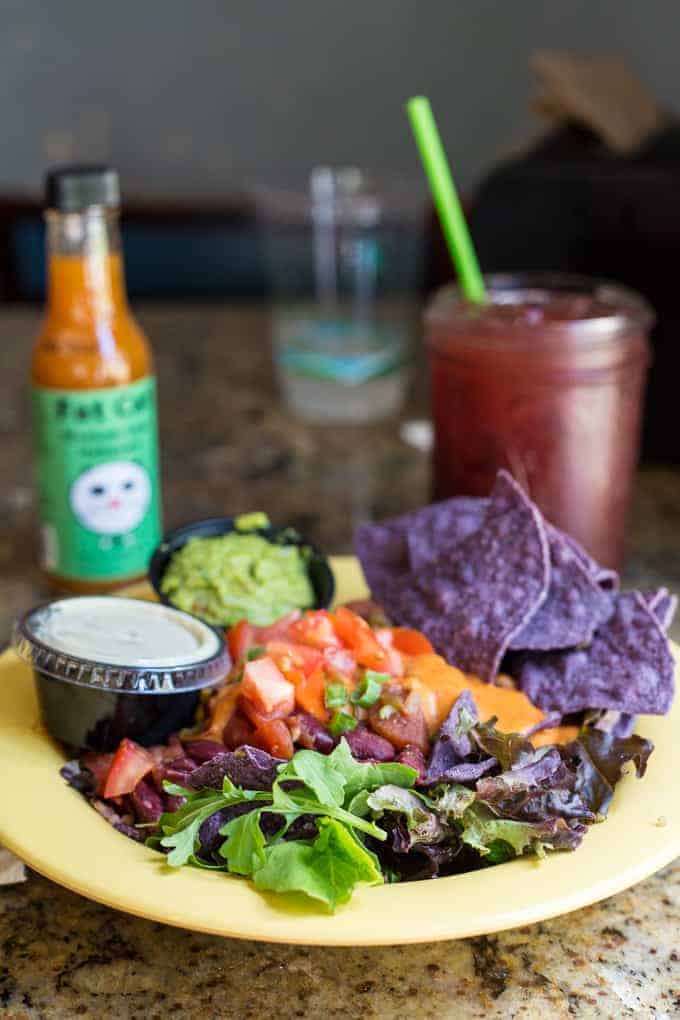 A yellow plate with mixed greens, diced tomatoes, beans, a small cup of dressing, guacamole, and blue corn tortilla chips. In the background, there is a bottle of hot sauce and a glass of dark red beverage with a green straw.