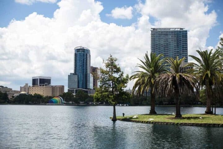 A cityscape with modern buildings and a rainbow-colored structure in the background, viewed across a lake with palm trees and a small island in the foreground under a partly cloudy sky.