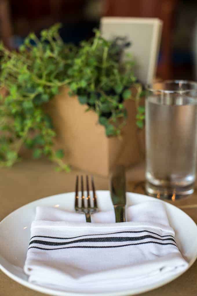 A white plate with a folded white napkin, fork, and knife sits on a table. A glass of water and a brown box planter with green leafy plants are in the background.