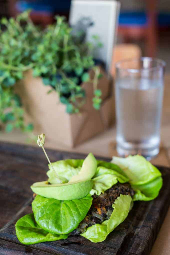 A lettuce-wrapped burger patty topped with avocado slices, held together by a toothpick, sits on a wooden board; a glass of water and a potted plant are in the background.
