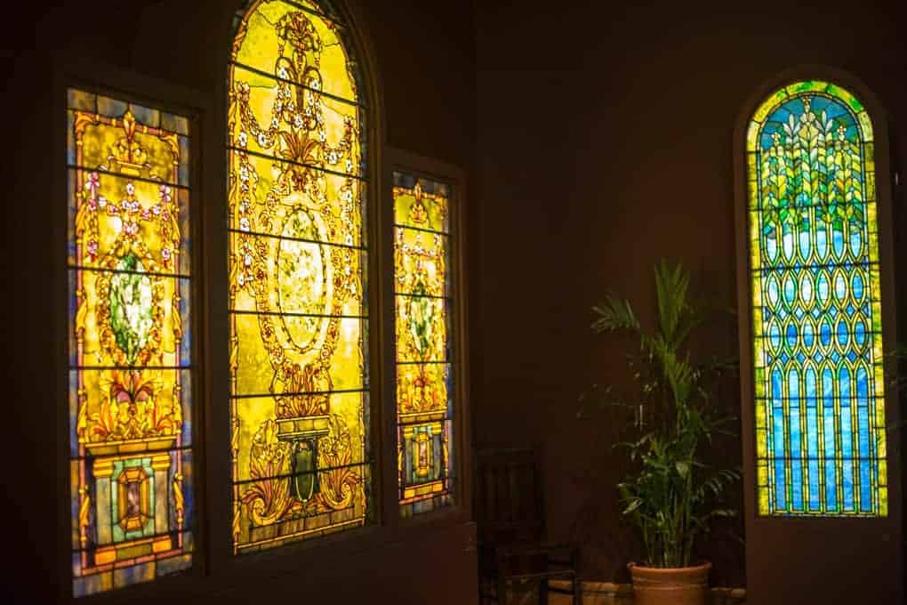 Three stained glass windows in a dimly lit room; two on the left feature intricate yellow and gold designs, while the one on the right has a blue and green geometric pattern. A potted plant stands below the windows.
