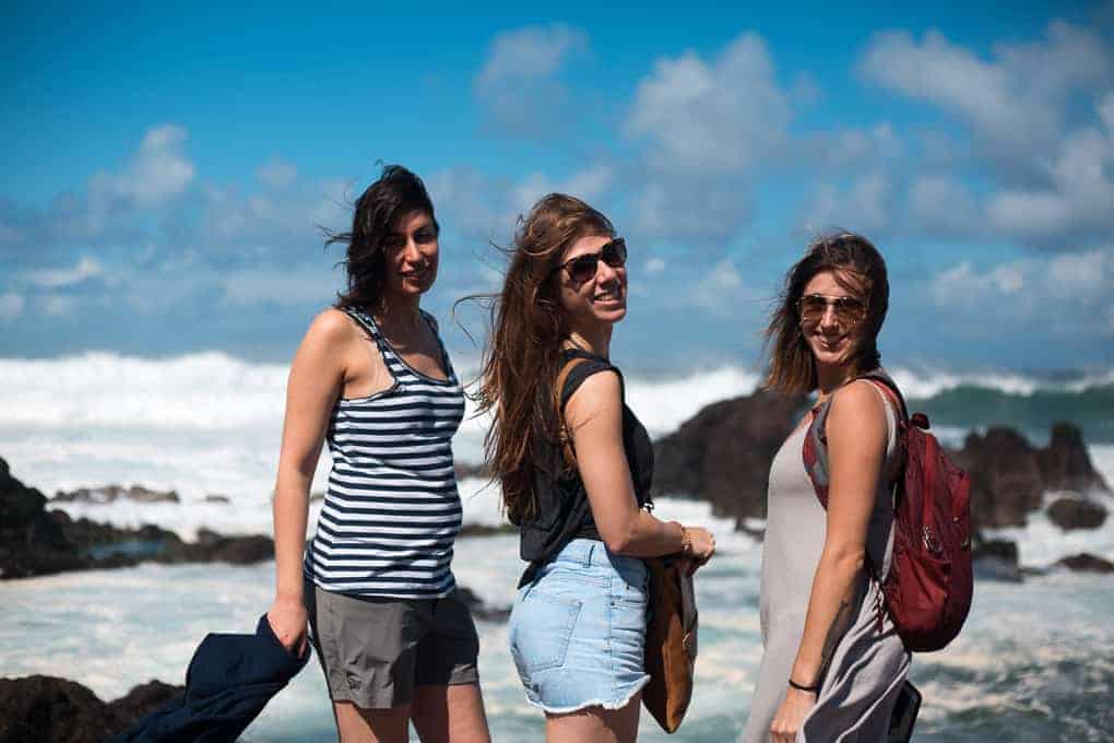 Three women stand outdoors by rocky ocean waves under a blue sky with clouds. They are facing the camera, wearing casual summer clothing, and one has a red backpack. The wind is blowing their hair.