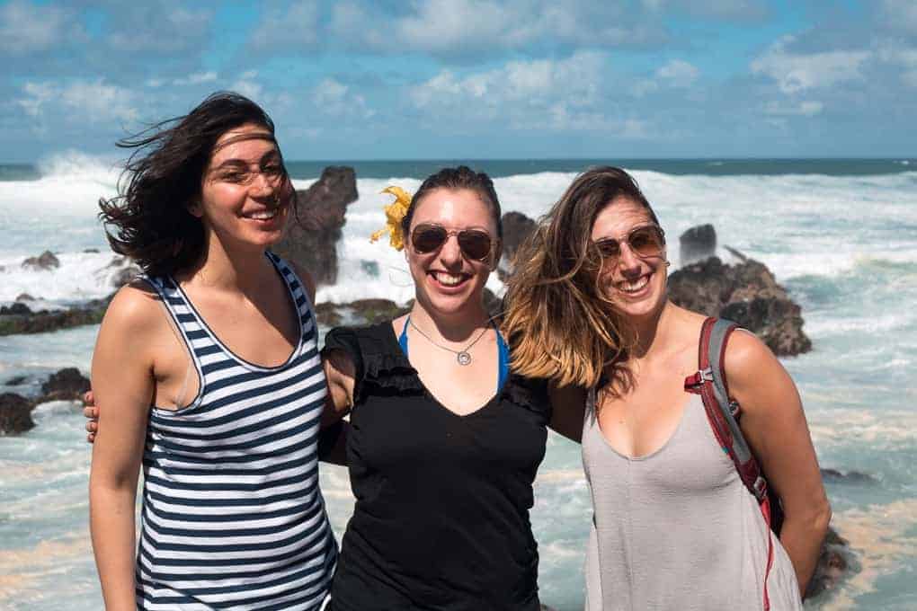 Three women stand together smiling in front of a rocky shoreline with waves crashing behind them. They are dressed in casual summer clothes and sunglasses, with bright daylight and a partly cloudy sky.
