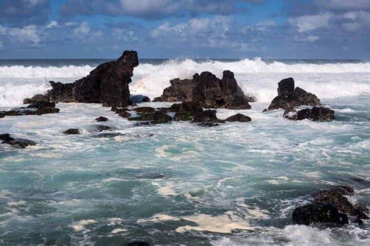 Dark, jagged rocks protrude from choppy, foamy ocean water under a partly cloudy sky. Waves crash against the rocks, creating white spray and swirling water in the foreground.