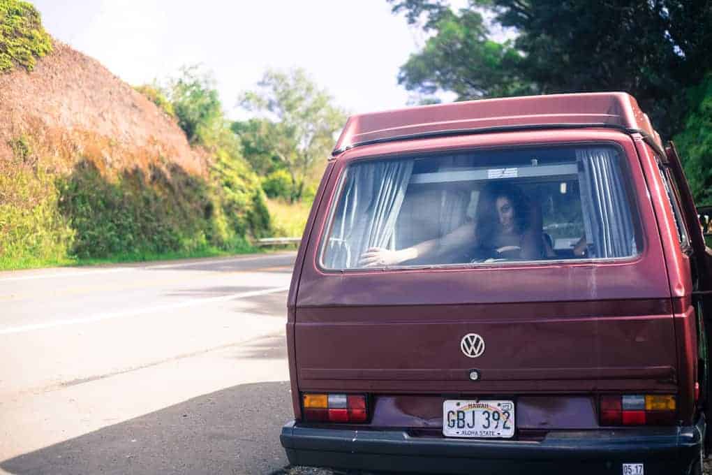 A maroon Volkswagen van with a visible license plate is parked by the side of a road. A person sits inside, partially visible through the rear window. Trees and greenery are in the background.