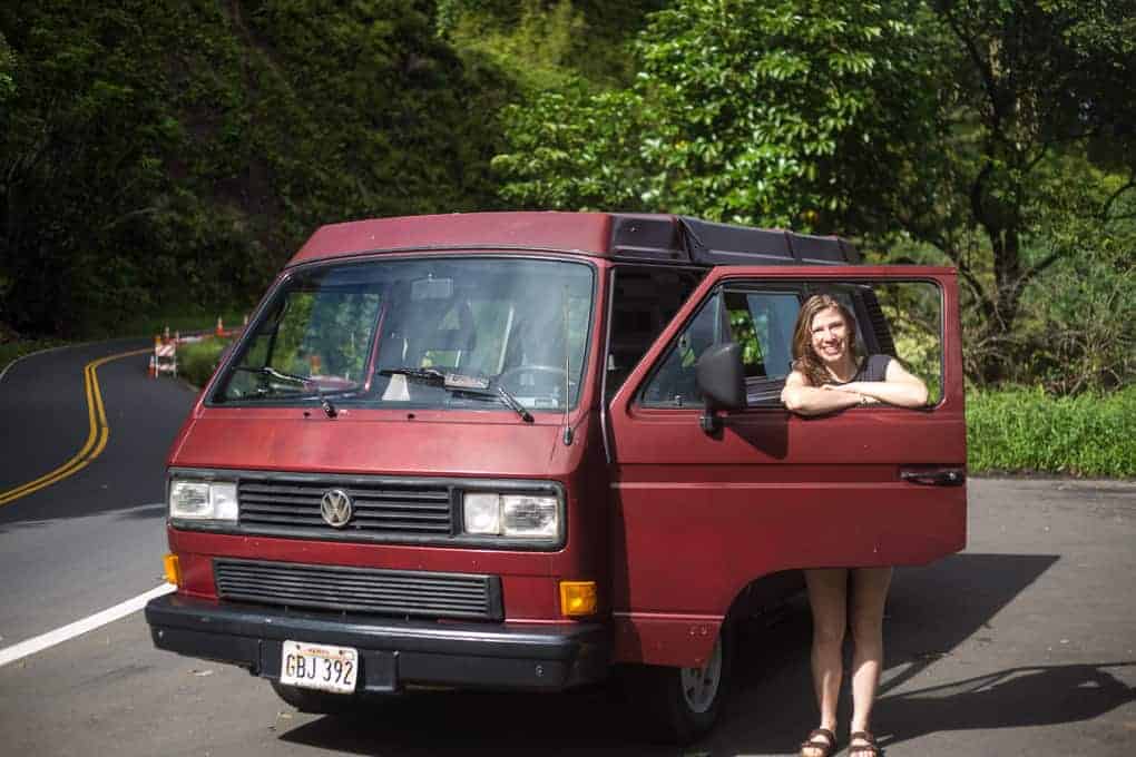 A person stands smiling with arms resting on the open door of a red Volkswagen van parked on the side of a curving road surrounded by greenery and trees.