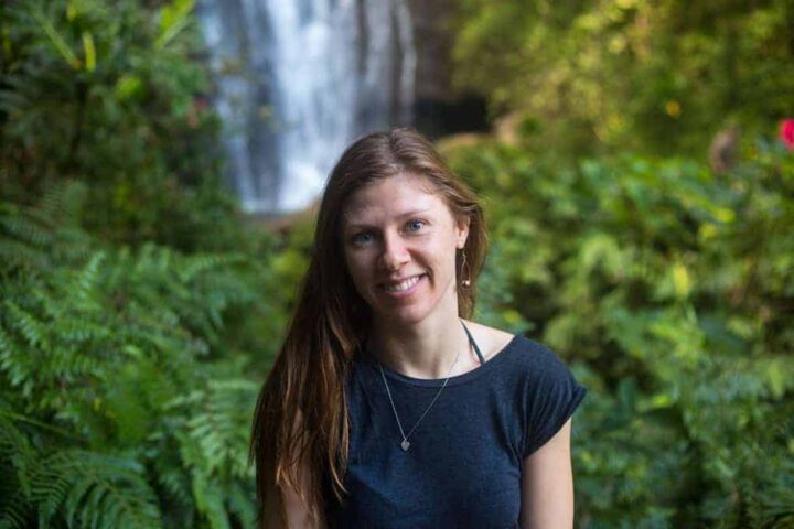 A woman with long brown hair and a black t-shirt stands outdoors in front of green foliage, with a waterfall visible in the blurred background.